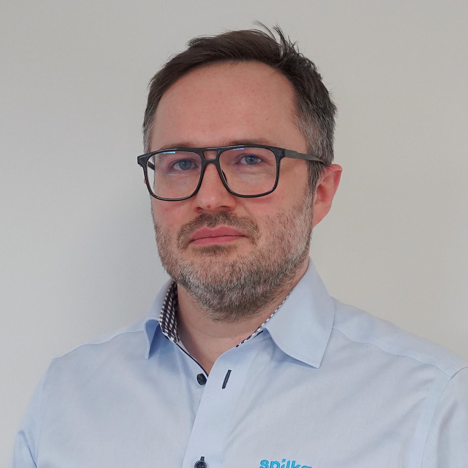 A middle-aged man with short dark hair, glasses, and a beard, wearing a light blue collared shirt with a patterned inner lining, standing against a plain white background.