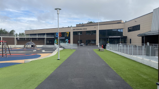 En skolebygning sett fra en asfaltvei, med lekeområde til venstre og grønt gress til høyre, under en skyet himmel.