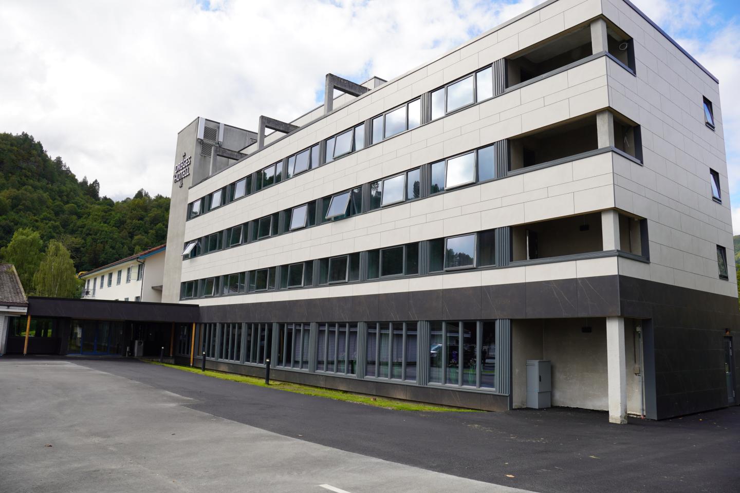 Moderne hotellbygg med flere etasjer, store vinduer og en lobbyinngang, omgitt av fjell og grønt landskap under en delvis skyet himmel.