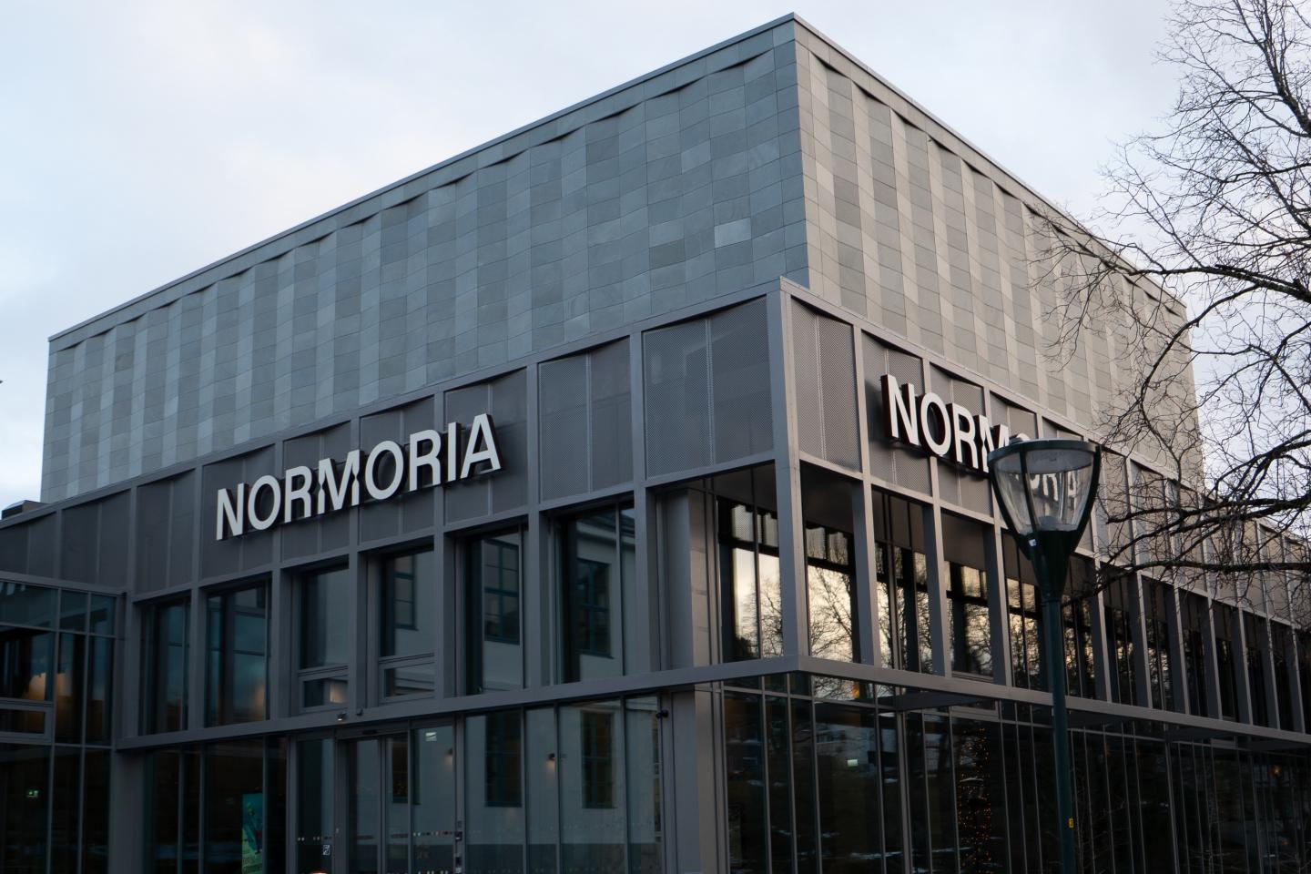 Et moderne bygning med store glassvinduer og skilt som sier "NORMORIA" på fasaden.
