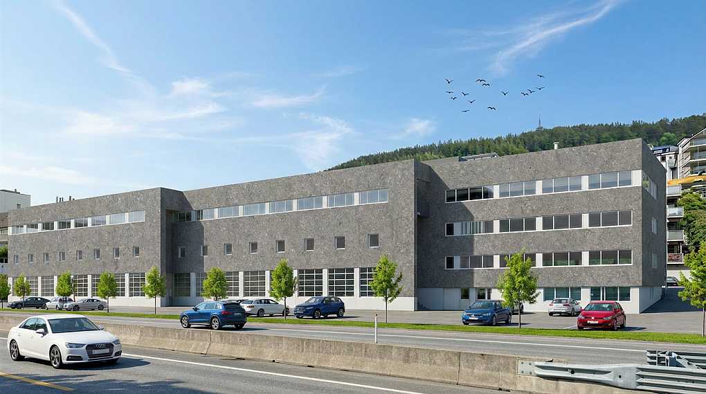 Modernt grå bygning med små og store vinduer, omgitt av parkeringsplasser og grønne trær, under en klar blå himmel.