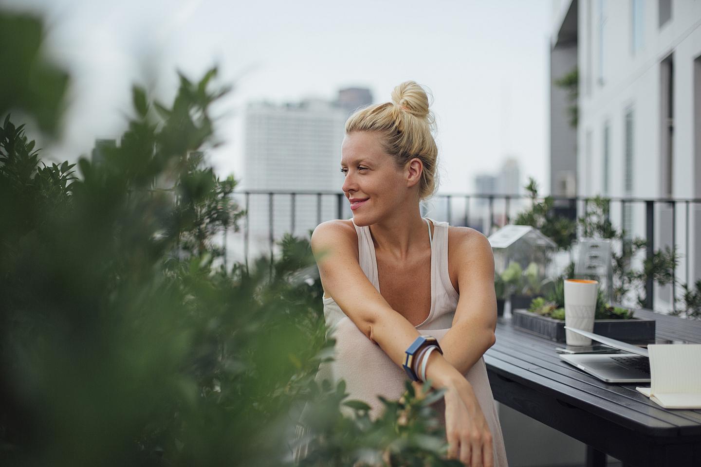 milende kvinne som jobber på en balkong med planter, bylandskap i bakgrunnen, med en laptop og notatbok på bordet